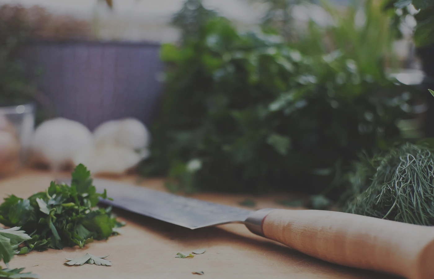 A chef's knife resting on a worn wooden board beside finely chopped parsley.