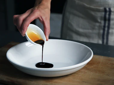 A hand pouring dark sauce from a small jug into a white bowl.