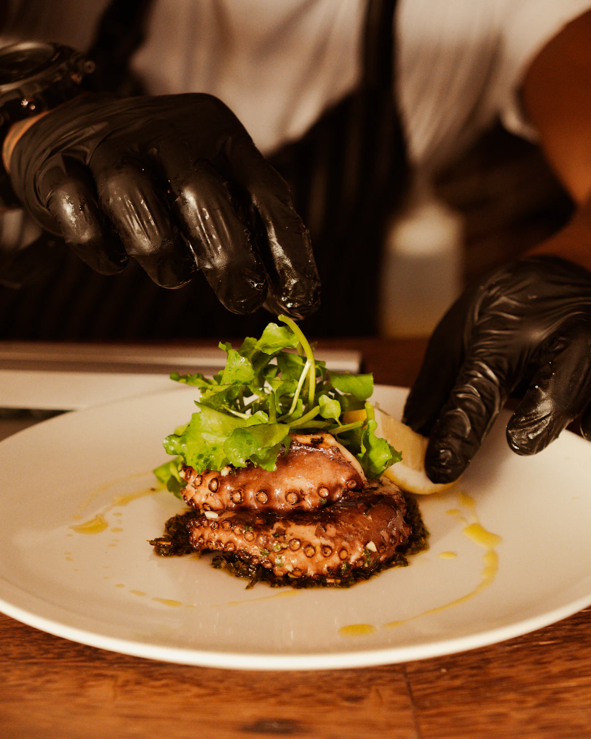 Gloved hands laying charred leek and greens in a low line across a wide ivory plate.
