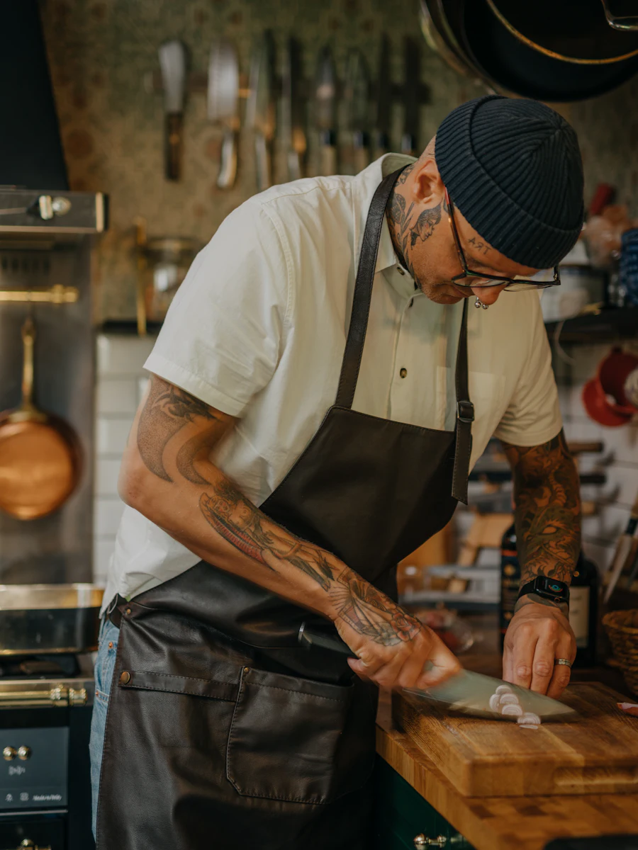Idris Kalfa, tattooed and in an apron, slicing garlic on a wooden board in the studio kitchen.
