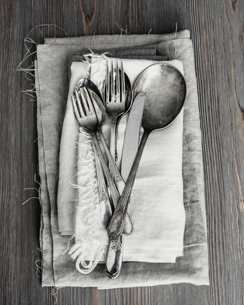 Worn silver fork, knife and spoon resting on frayed linen napkins over old wood.