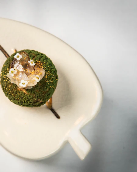 Cod under a bright herb crumb with small white flowers, served on a leaf-shaped dish.